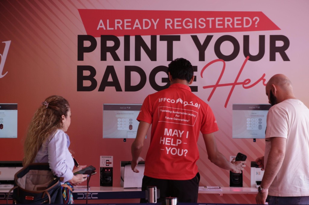 Event staff in a red uniform assisting attendees at Gulfood self-service badge printing stations